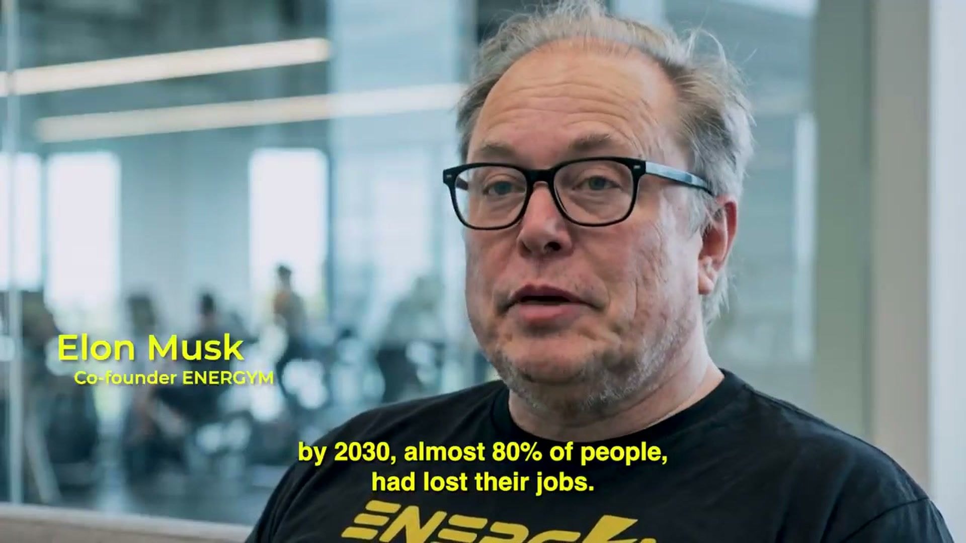 A greying, older Elon Musk in an Energym t-shirt, labelled Co-founder ENERGYM, being interviewed in a gym in the mockumentary