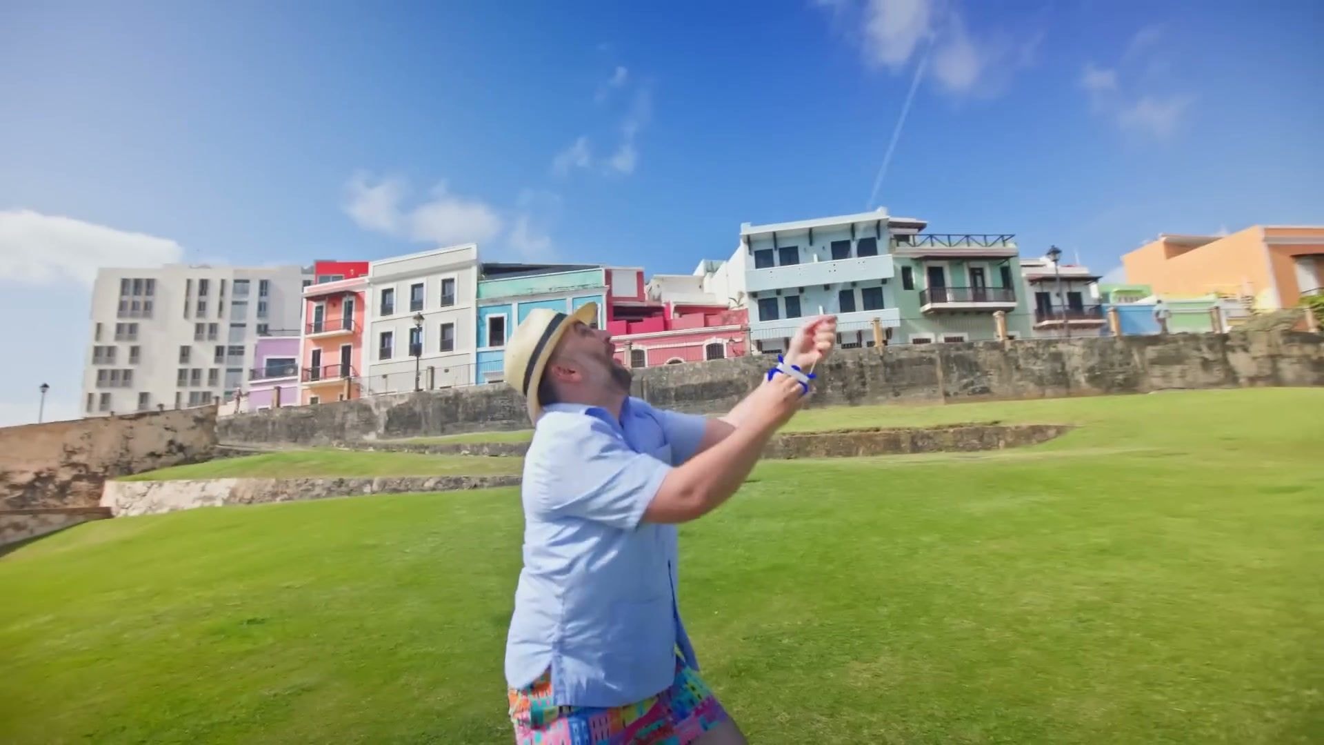 Bill Stiteler in a straw hat and blue shirt flying a kite on the grass below the city wall, with the coloured houses of Old San Juan behind him