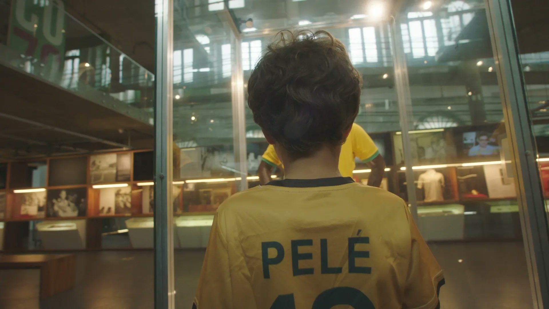 Seen from behind, a boy in a yellow shirt with PELÉ printed across the shoulders stands close to a tall glass display case in a museum hall, lit exhibition cabinets running along the walls behind him